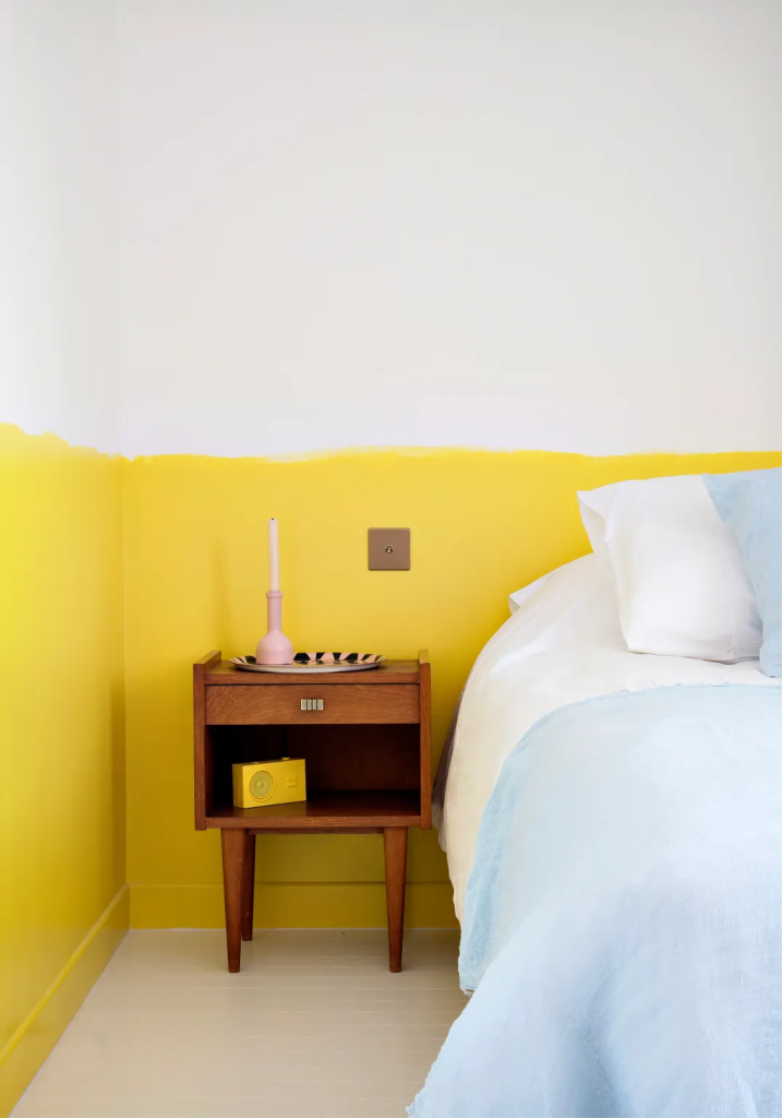 A bed next to a brown nightstand in the corner of a bedroom that is painted unevenly to about a quarter of the way up the wall.