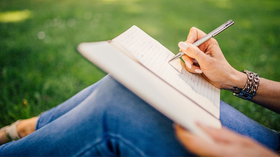 Person journaling in a notebook outdoors on the grass, promoting mindfulness and self-reflection.