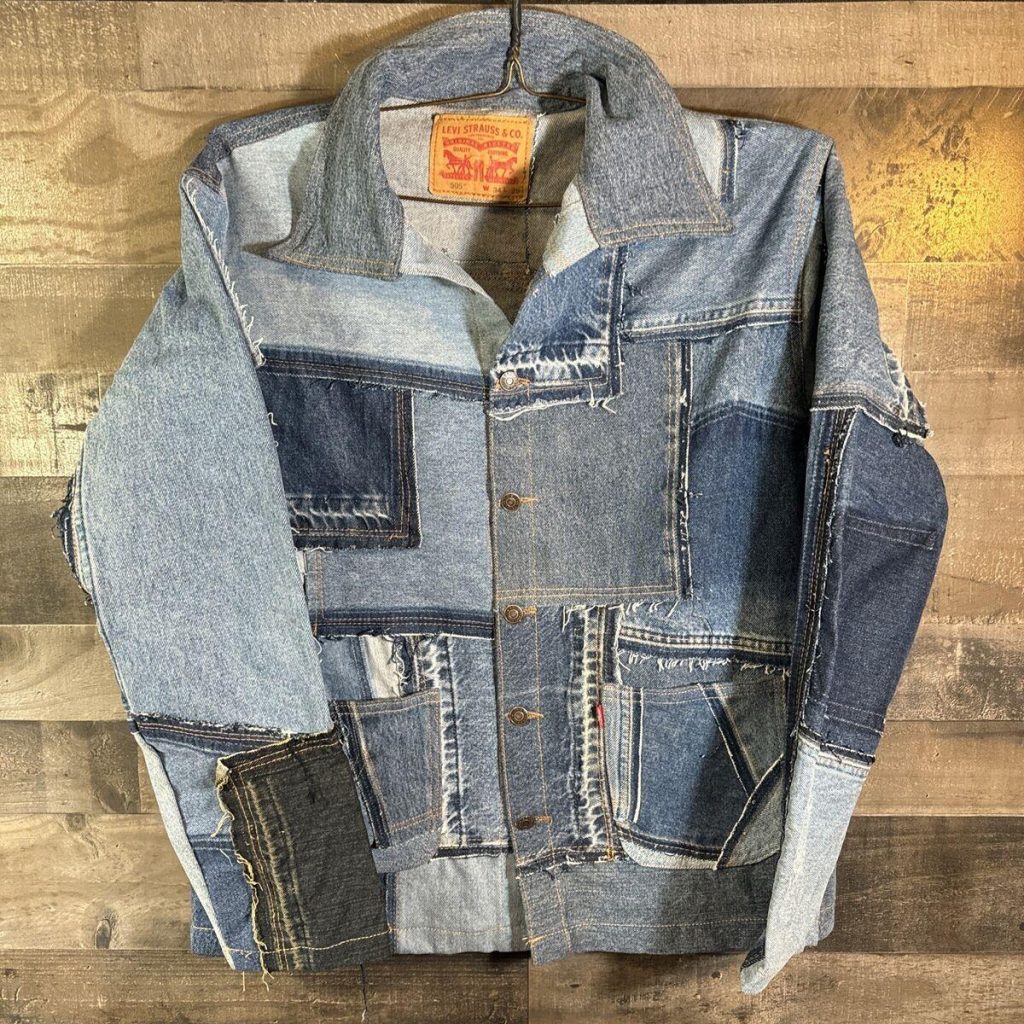 A stylish patchwork denim jacket made from various shades and pieces of recycled jeans, hung on a rustic wooden wall.