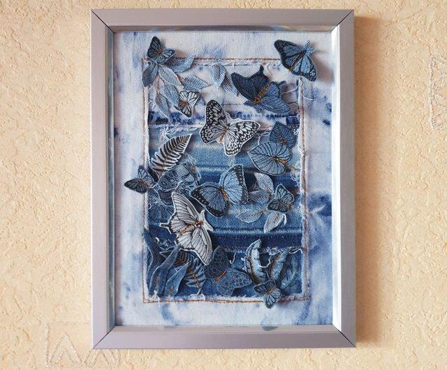 A framed wall art piece featuring intricately cut and sewn denim butterflies and foliage, arranged on a tie-dye blue denim background.
