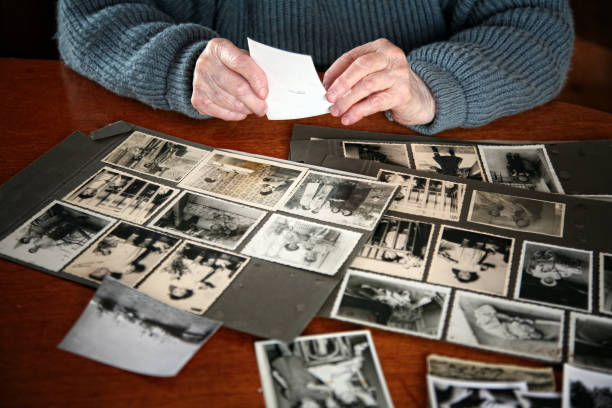 Black and white photos laying on a table most stuck to black canvas paper with a seated elderly individual sorting through each photo.