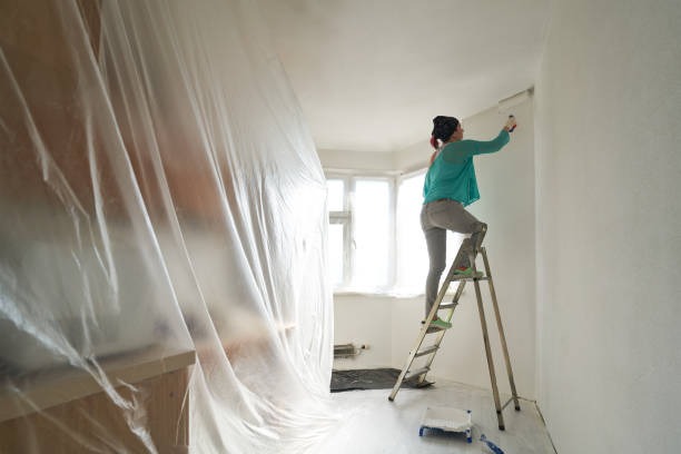 A woman on a ladder applying paint while appliances are covered.