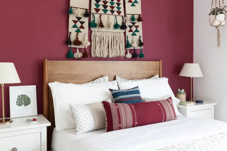 A bed against a maroon colored accent wall with white nightstands on either side and plenty of pillows on the bed.