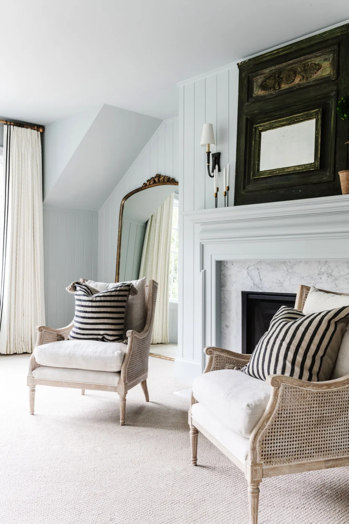 A mirror reflecting light from a window in the corner of a bedroom behind seating in front of a modern fireplace..