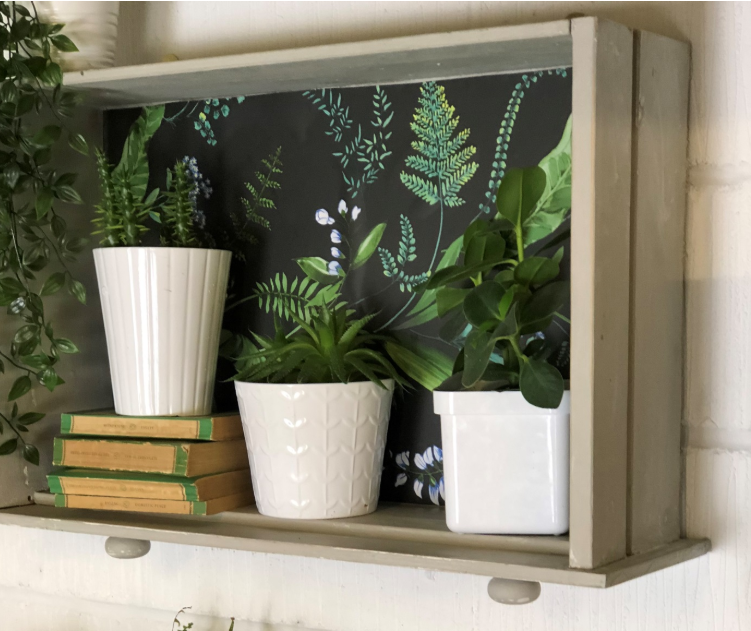An old white drawer affixed to the white wall by its bottom while being used as a shelf holding potted plants and a few old books.