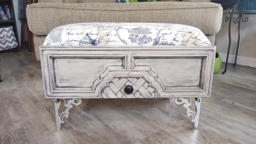 A antique looking white drawer fitted with mirrored intricately designed legs with a cushion affied making it a cozy foot rest.