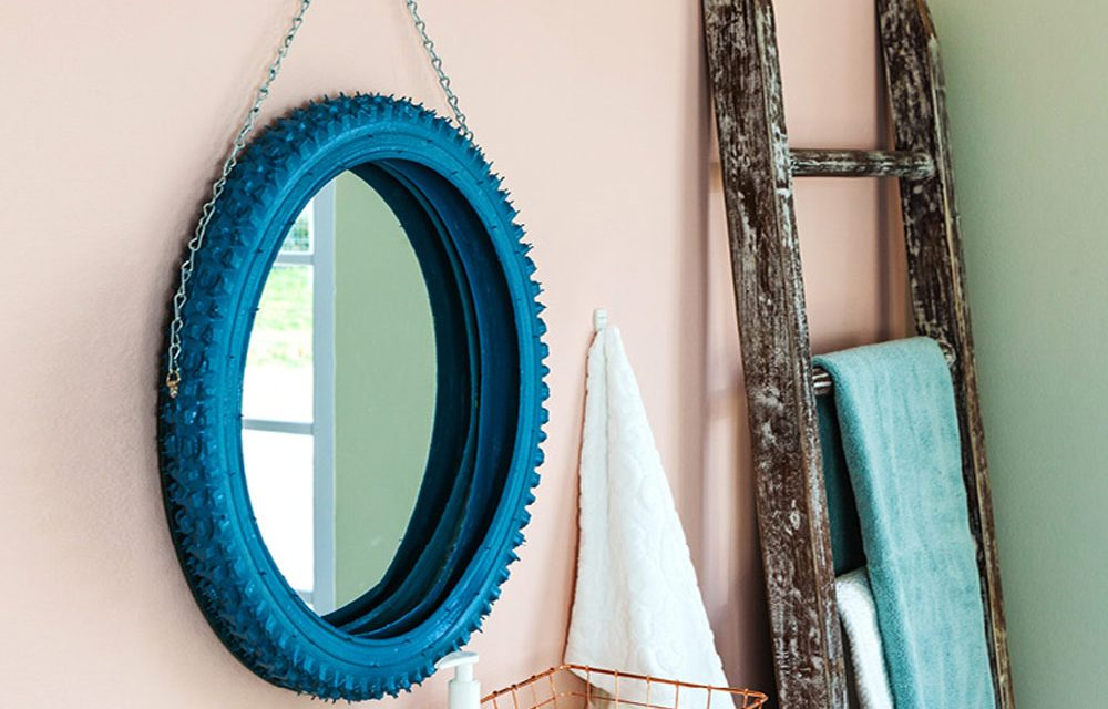 Bicycle tire painted blue hanging form a chain on a peach colored bathroom wall with a mirror inside.