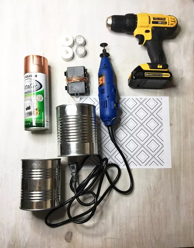 Top down view of two tin cans, two tea lights, a can of spray paint, and a drill on a tabletop.