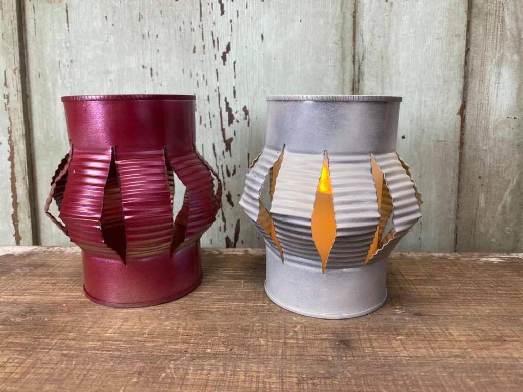 Two tin can lanterns on a table in front of a wooden wall designed with the center of the red and white cans sliced.