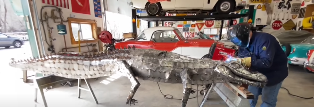 A beautiful crocodile made by Mike Orr in the foreground of a mechanic garage filled with classic cars in the background.