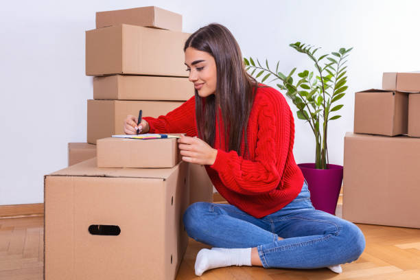 A woman in a red sweater and blue jeans sitting legs cross crossed on the floor surrounded by packed boxes and a potted plant but journaling on a notepad resting on a pile of boxes next to her with a smile.