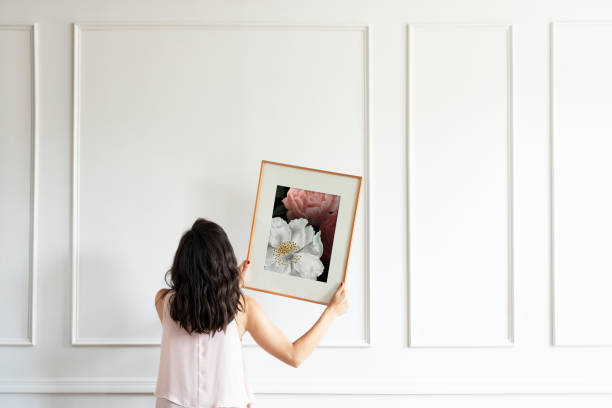 A woman about to place a framed painting of flowers on a white decorative wall.