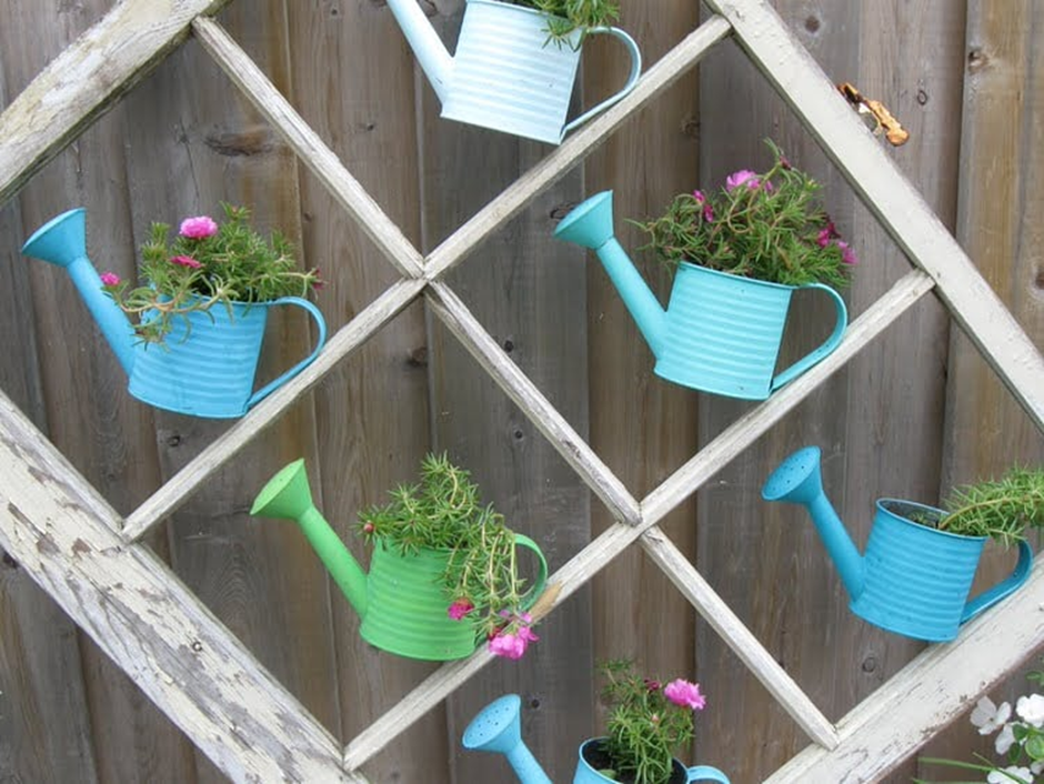 Old window frame used as a wall trellis with colorful watering cans filled with flowers.