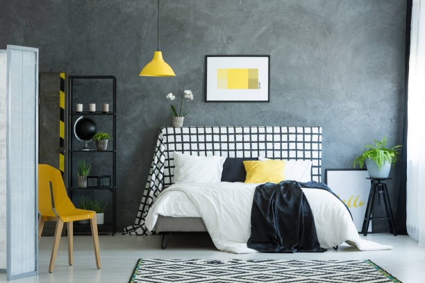 A modern industrial-style bedroom featuring a textured gray wall, black grid-pattern headboard, white bedding with yellow accents, and minimalist decor.