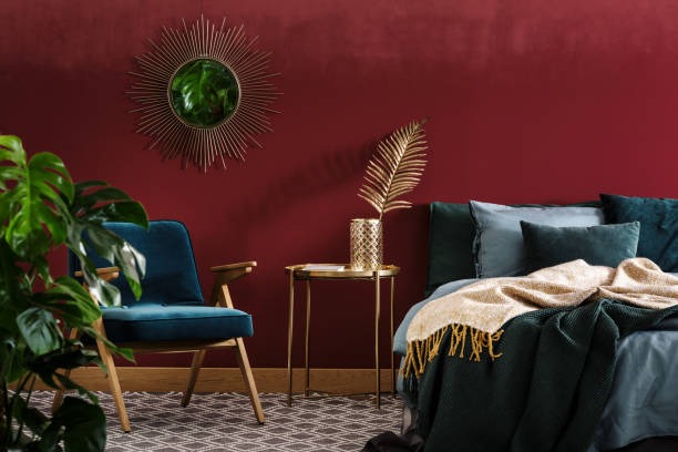 A stylish glam bedroom with a deep burgundy accent wall, velvet teal armchair, gold side table, and a sunburst mirror above a bed with dark green and gold bedding.