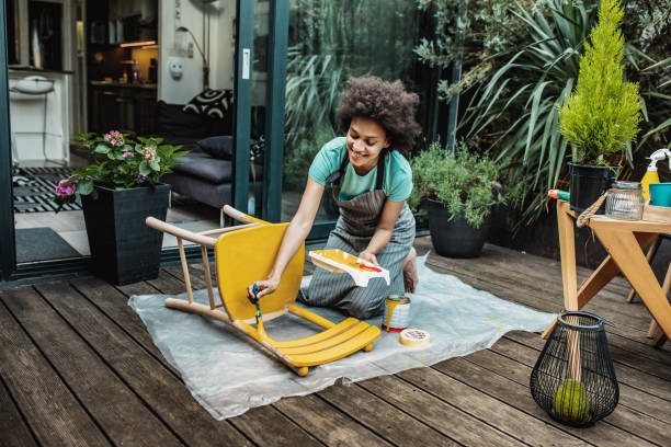 A woman on a outside deck painting a chair yellow.