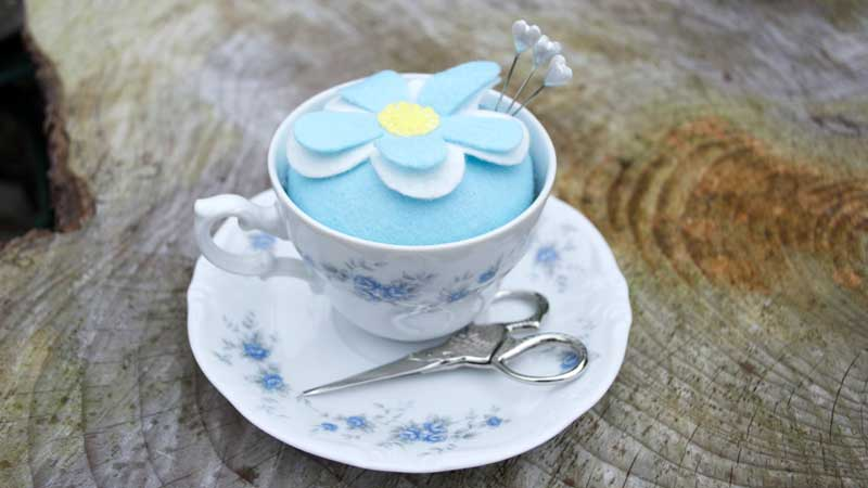 A teacup being used to hold a decorative baby blue pin cushion.