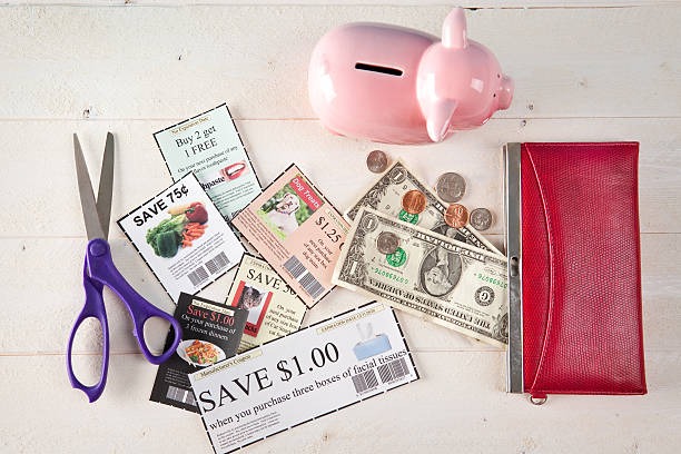 Top down view of coupons, a red purse, scissors, loose change, a dollar bill, and a pink piggy bank on a wooden backing.