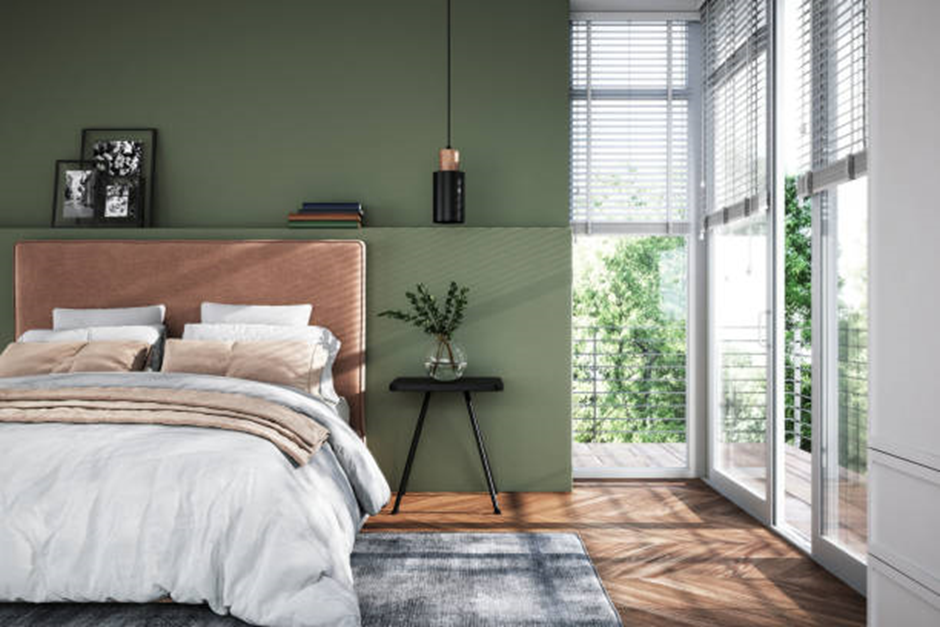 A contemporary bedroom featuring a muted green accent wall, a plush upholstered headboard, wood floors, and large windows with blinds overlooking a balcony.