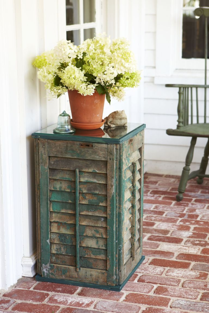 Rustic upcycled shutter cabinet with distressed green paint used as a porch accent table topped with a terracotta pot of white hydrangeas