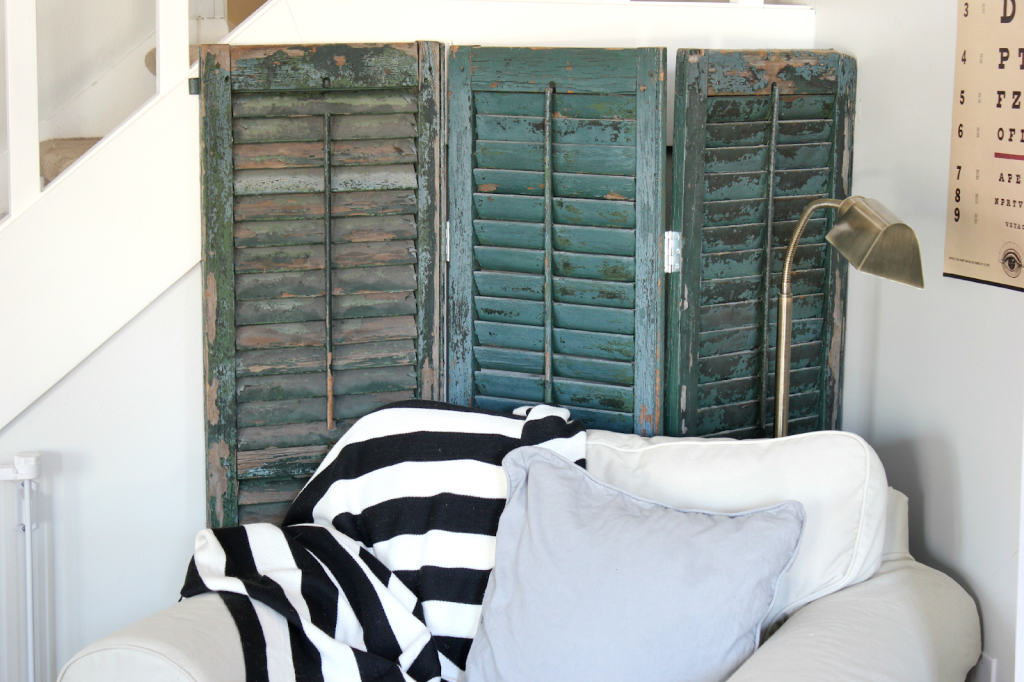 Vintage green distressed shutters repurposed as a decorative room divider placed behind a white sofa with striped blanket and pillow