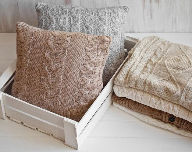 Decorative pillows made from repurposed cable-knit sweaters placed in a white wooden crate beside folded neutral-colored knit blankets.