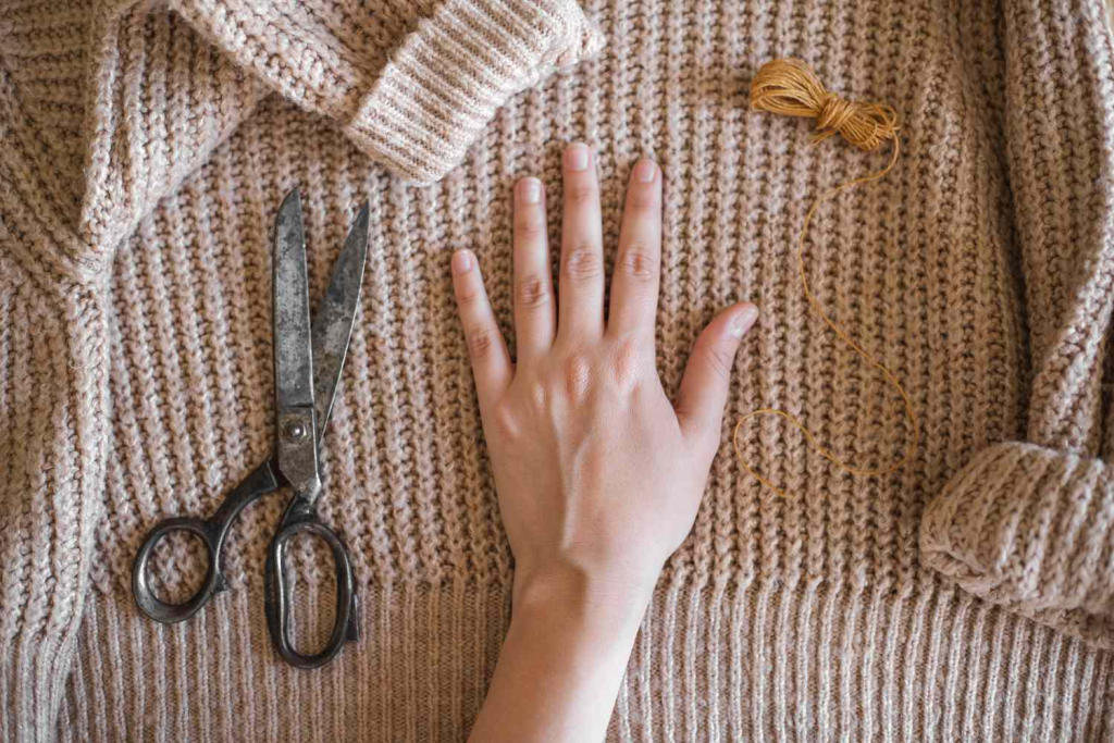 A hand laid flat on a knitted sweater next to scissors and yarn, preparing the sweater for an upcycling craft project.