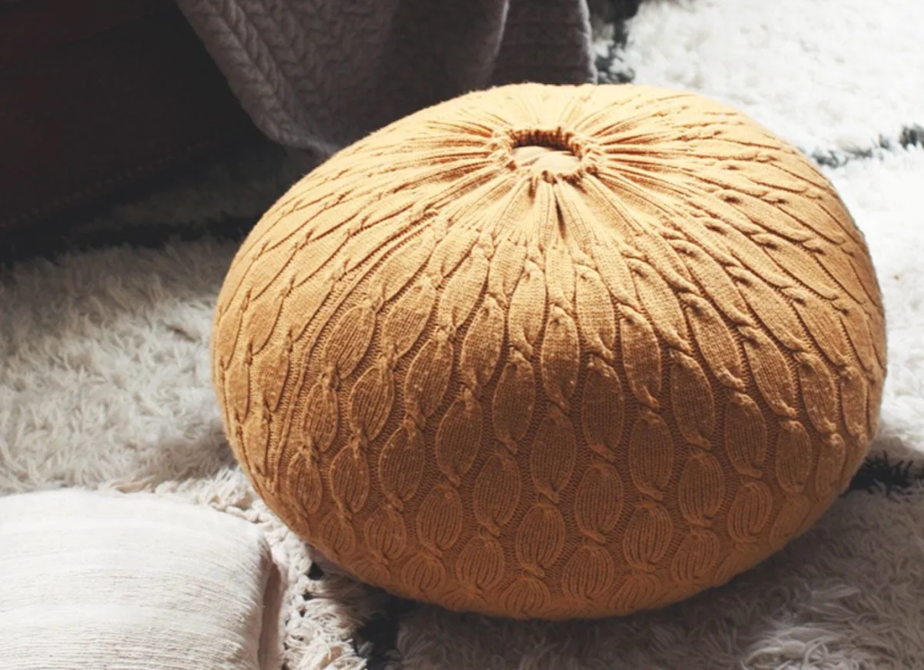 A cozy round pouf covered in mustard-colored cable-knit sweater fabric placed on a soft rug in a living room.