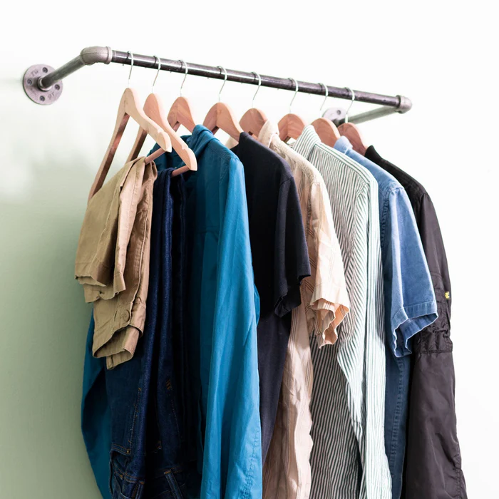 Wall-mounted industrial pipe clothing rack holding shirts and pants on wooden hangers.