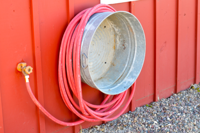 Upcycled metal bucket doubling as a smart garden hose holder affixed to the red slatted wooden siding of a storage shed.