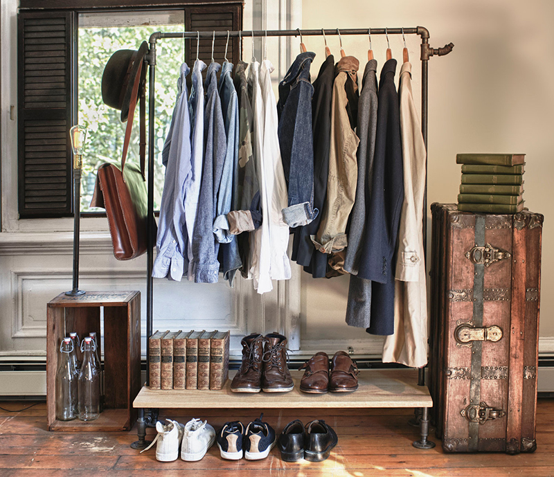 Industrial pipe clothing rack with shirts, jackets, shoes, books, and vintage storage trunks in a rustic room