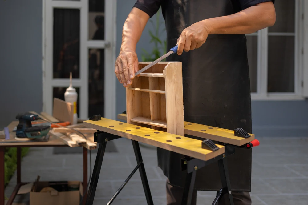 Woodworker filing a small wooden shelf unit on a portable workbench during DIY furniture build