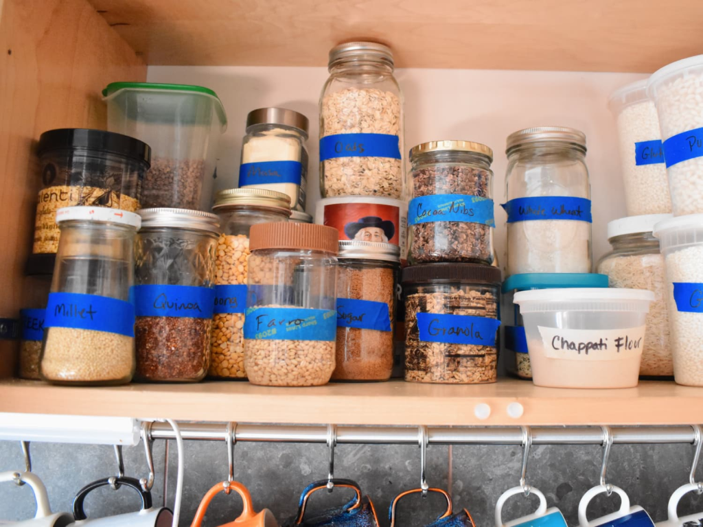 Labeled glass jars with oats, quinoa, flour, and pantry staples arranged neatly on kitchen shelf