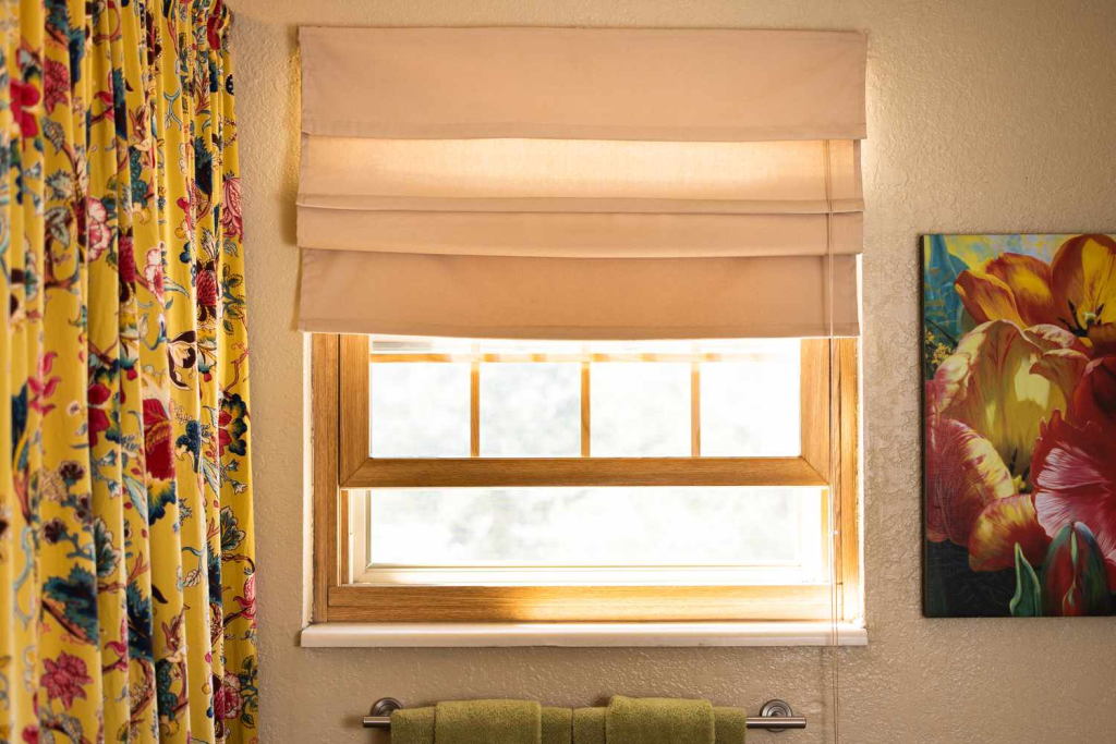 Bathroom window with beige roman shade and wooden frame, styled with floral curtain and wall art for a cozy interior look
