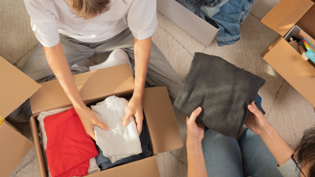 people packing folded clothes into cardboard box for moving or donation