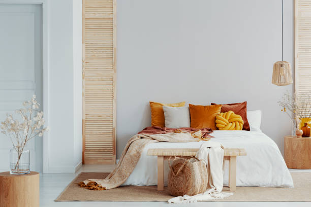Brown and orange pillows on white bed in natural bedroom interior with wicker lamp and wooden bedside table with vase.