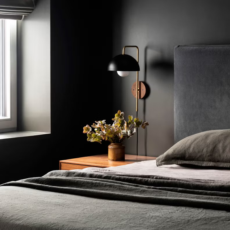 Dark bedroom corner with gray bedding and floral decor on nightstand.