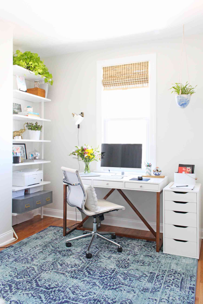 Bright home office with a white desk, rolling chair, blue patterned rug, and wall shelves with plants and decor.