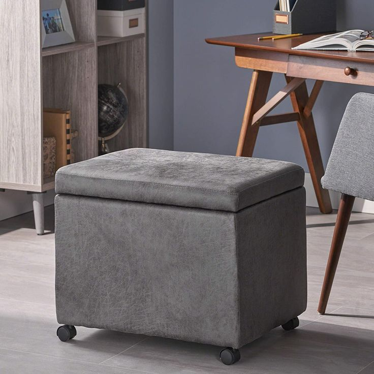 Compact grey ottoman with storage on wheels placed next to a mid-century wooden desk and grey chair.