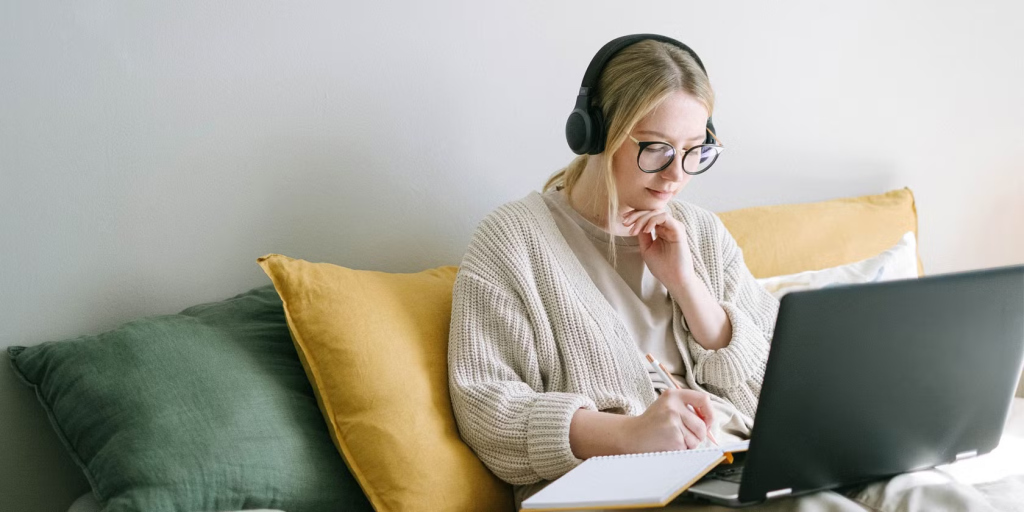 Woman using laptop and writing in notebook on couch.