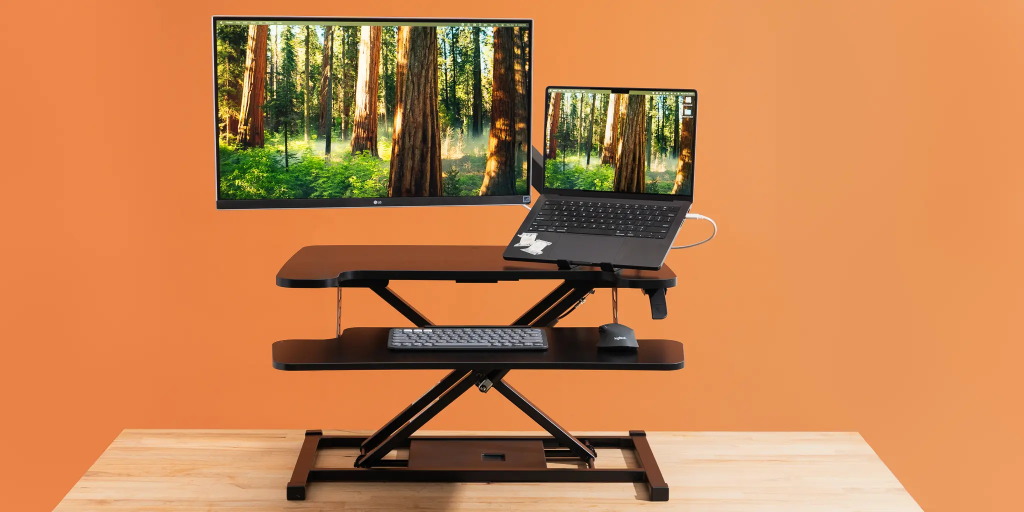 Adjustable standing desk with a dual monitor and laptop setup against an orange wall.