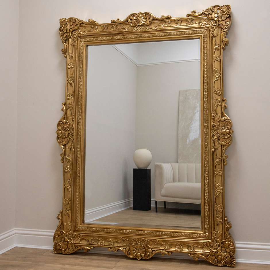 Ornate gold baroque floor mirror leaning against a wall in a minimalist cream space.