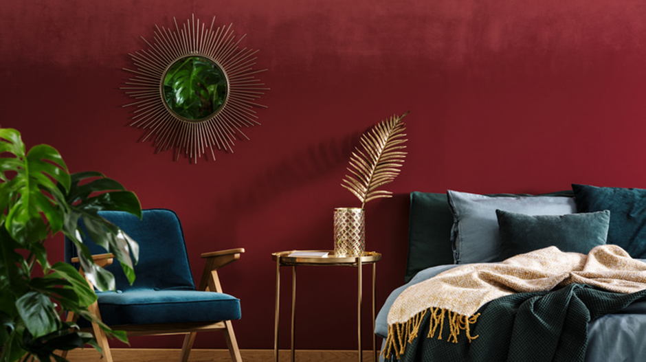 Stylish bedroom with burgundy wall, gold sunburst mirror, velvet teal chair, and cozy layered bedding.