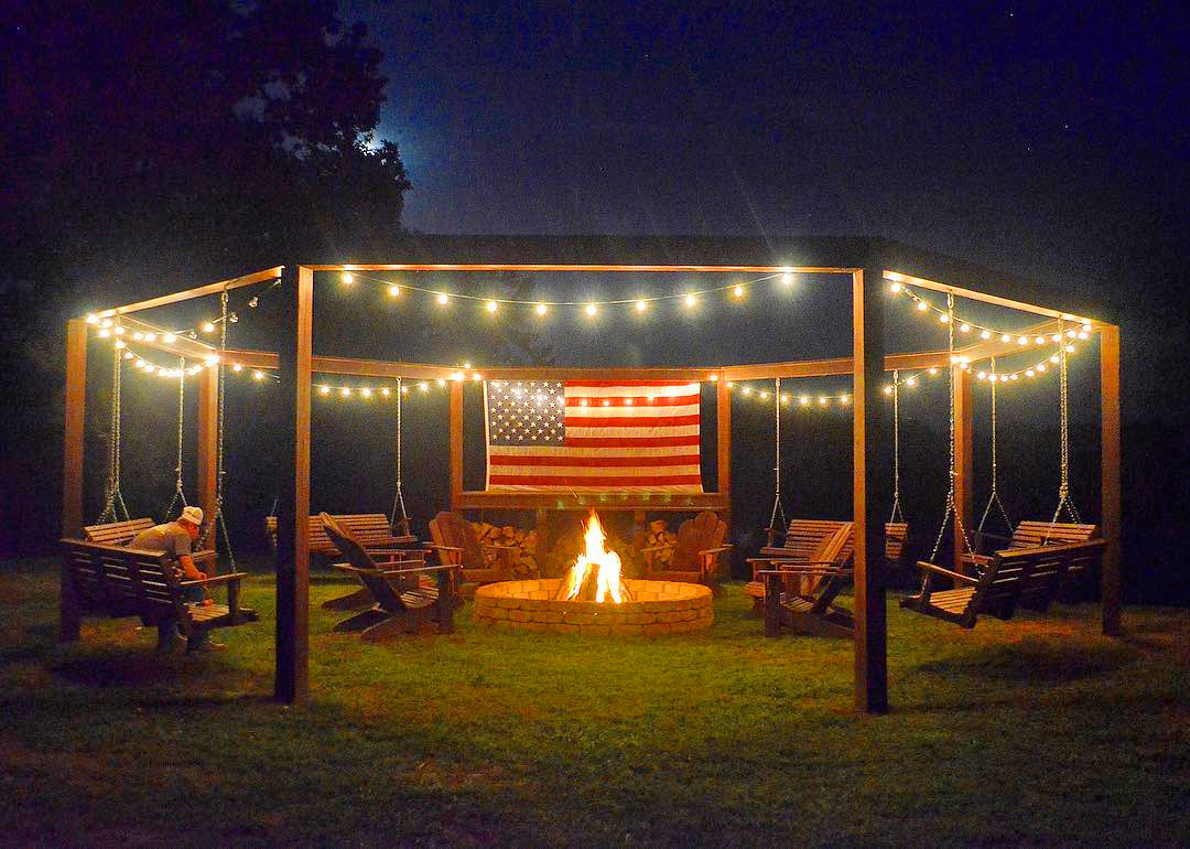 A pergola decorated with string lights surrounding a fire pit chairs and in the center an American flag.
