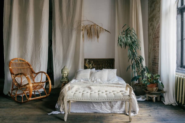 Boho bedroom with white bedding, rattan chair, indoor plants, and flowing curtains
